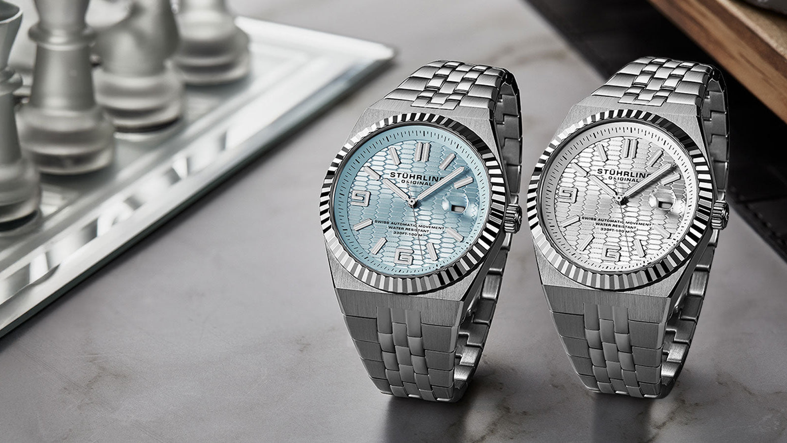 Two silver watches with detailed faces on a reflective surface next to a chessboard.
