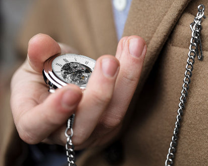 Hand holding a pocket watch with a chain, wearing a brown coat.