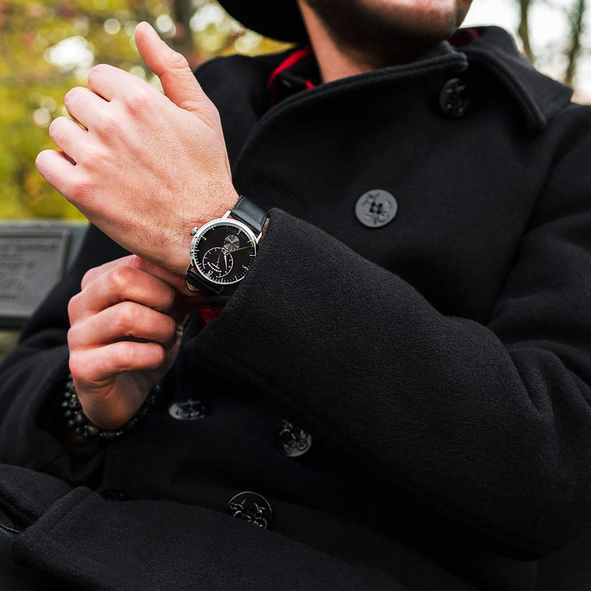 Person wearing a black coat and a watch, with a blurred outdoor background
